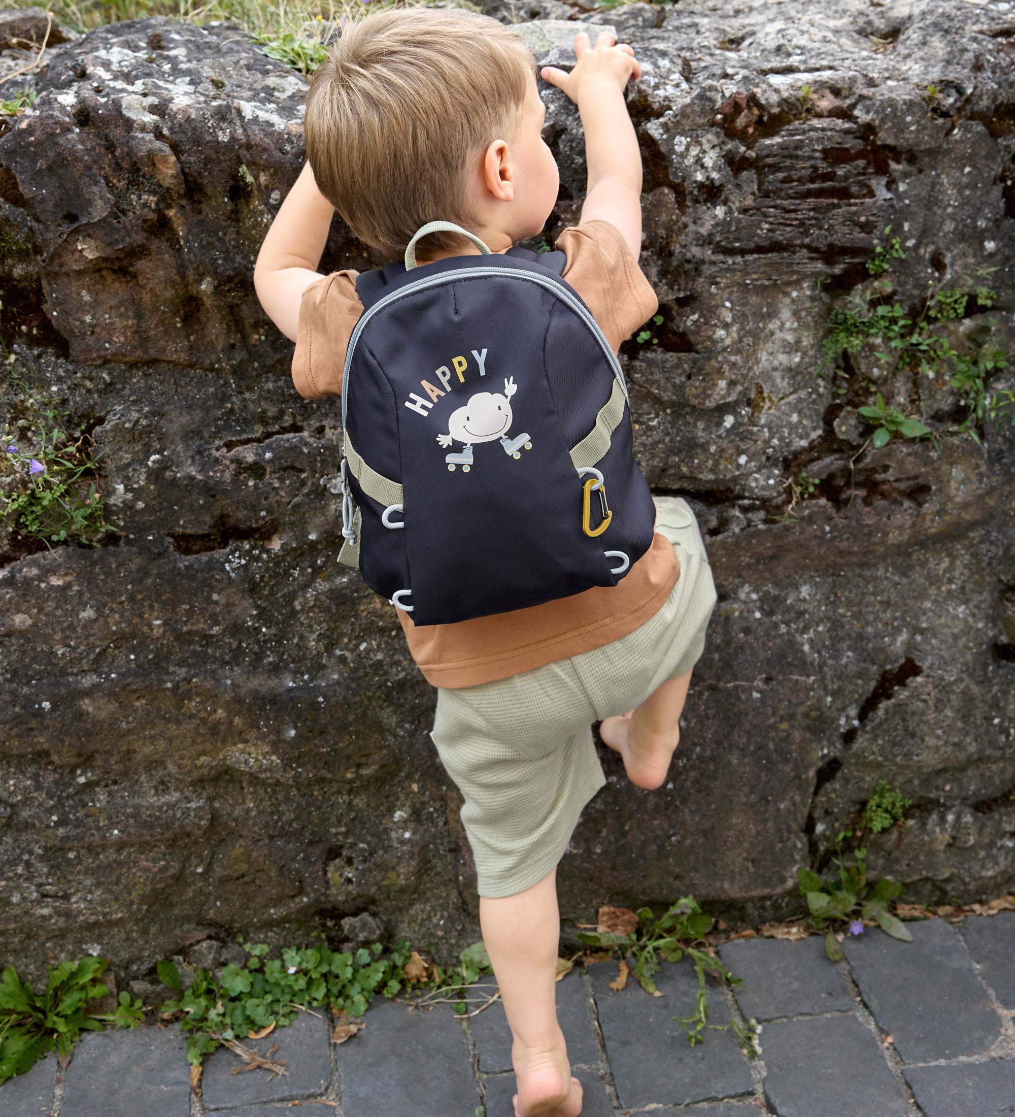 A little boy climbing a wall, wearing the Lassig tiny kids backpack for toddlers and little children, in a dark navy blue.