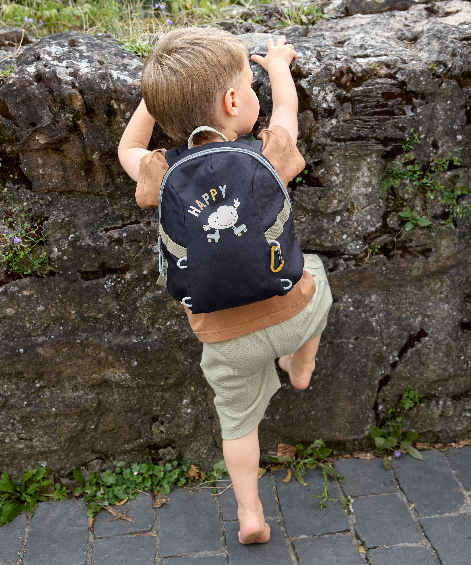 A little boy climbing a wall, wearing the Lassig tiny kids backpack for toddlers and little children, in a dark navy blue.