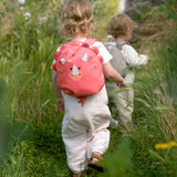 A little child walking through a field, wearing the Lassig Kids Tiny Backpack in a bright coral colour with a dinosaur face and 3D dino features.