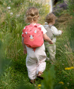 A little child walking through a field, wearing the Lassig Kids Tiny Backpack in a bright coral colour with a dinosaur face and 3D dino features.