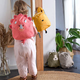 A child looking in the mirror, and wearing the Lassig Kids Tiny Backpack in a bright coral colour with a dinosaur face and 3D dino features.