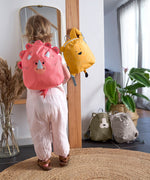 A child looking in the mirror, and wearing the Lassig Kids Tiny Backpack in a bright coral colour with a dinosaur face and 3D dino features.