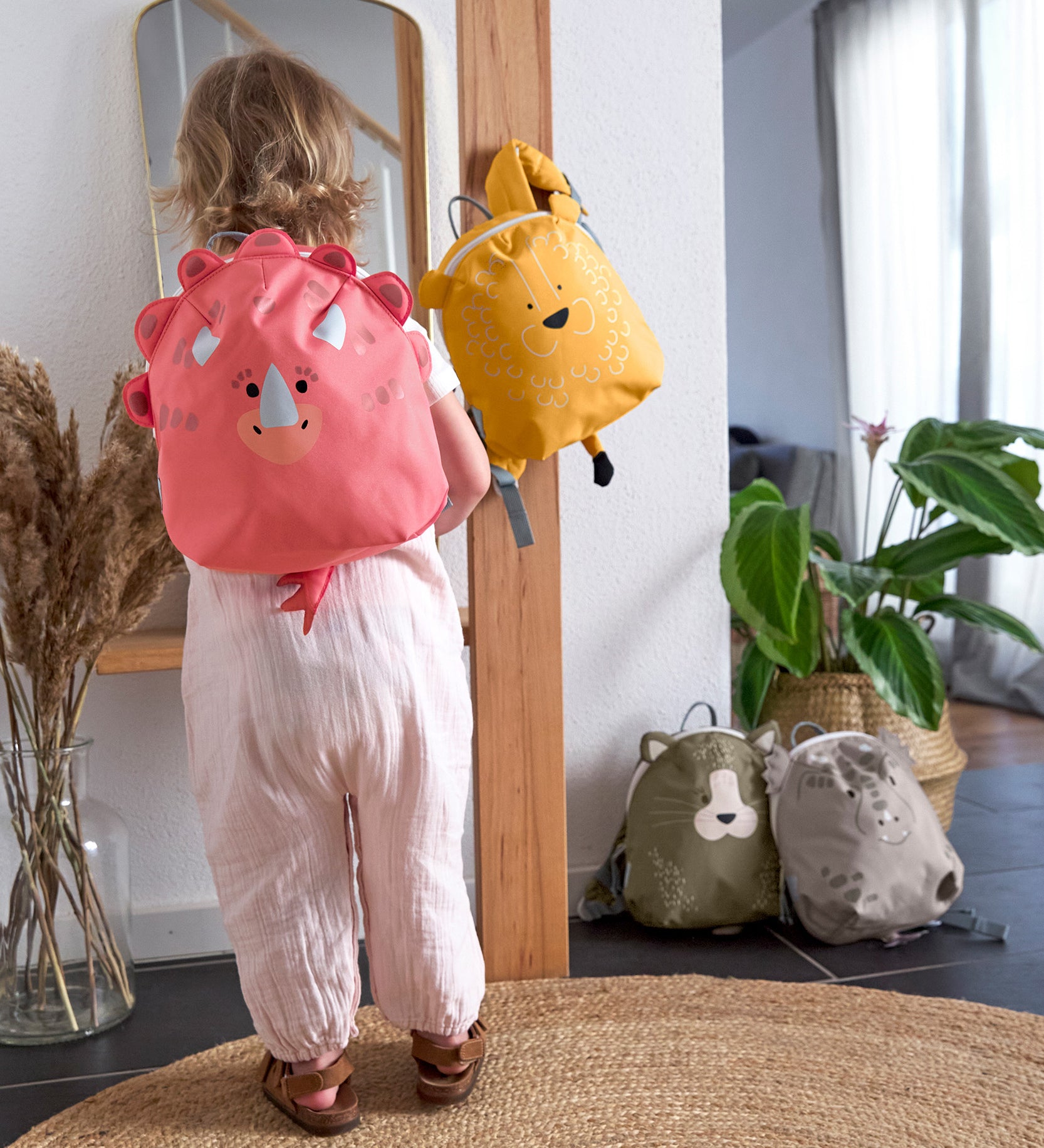 A child looking in the mirror, and wearing the Lassig Kids Tiny Backpack in a bright coral colour with a dinosaur face and 3D dino features.
