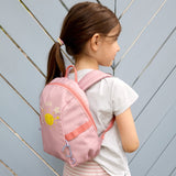 A girl wearing the Lassig tiny backpack for toddlers and little kids, in a light pink with a sunshine face cartoon.