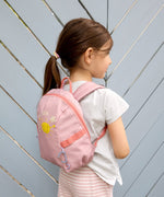 A girl wearing the Lassig tiny backpack for toddlers and little kids, in a light pink with a sunshine face cartoon.