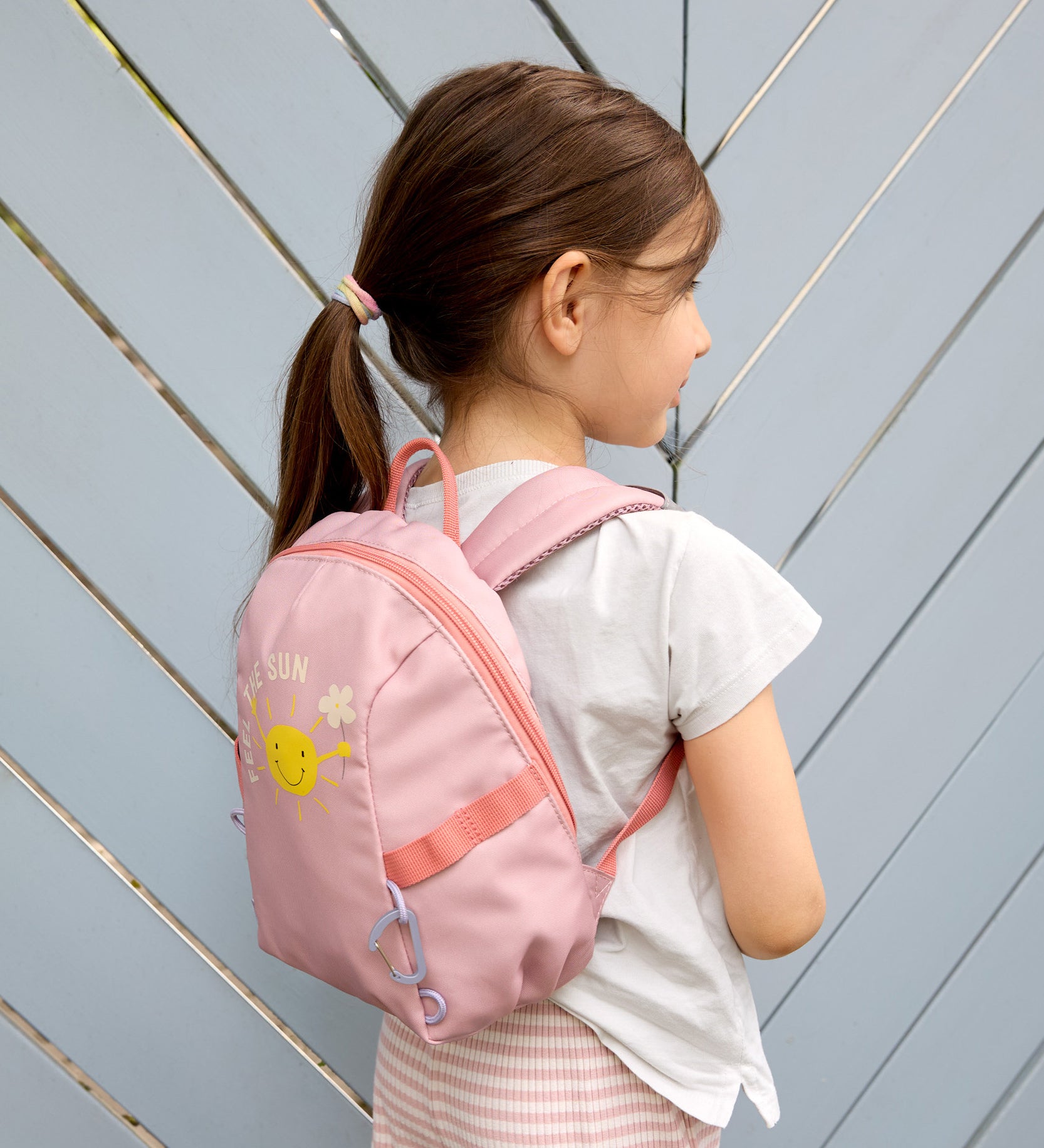 A girl wearing the Lassig tiny backpack for toddlers and little kids, in a light pink with a sunshine face cartoon.