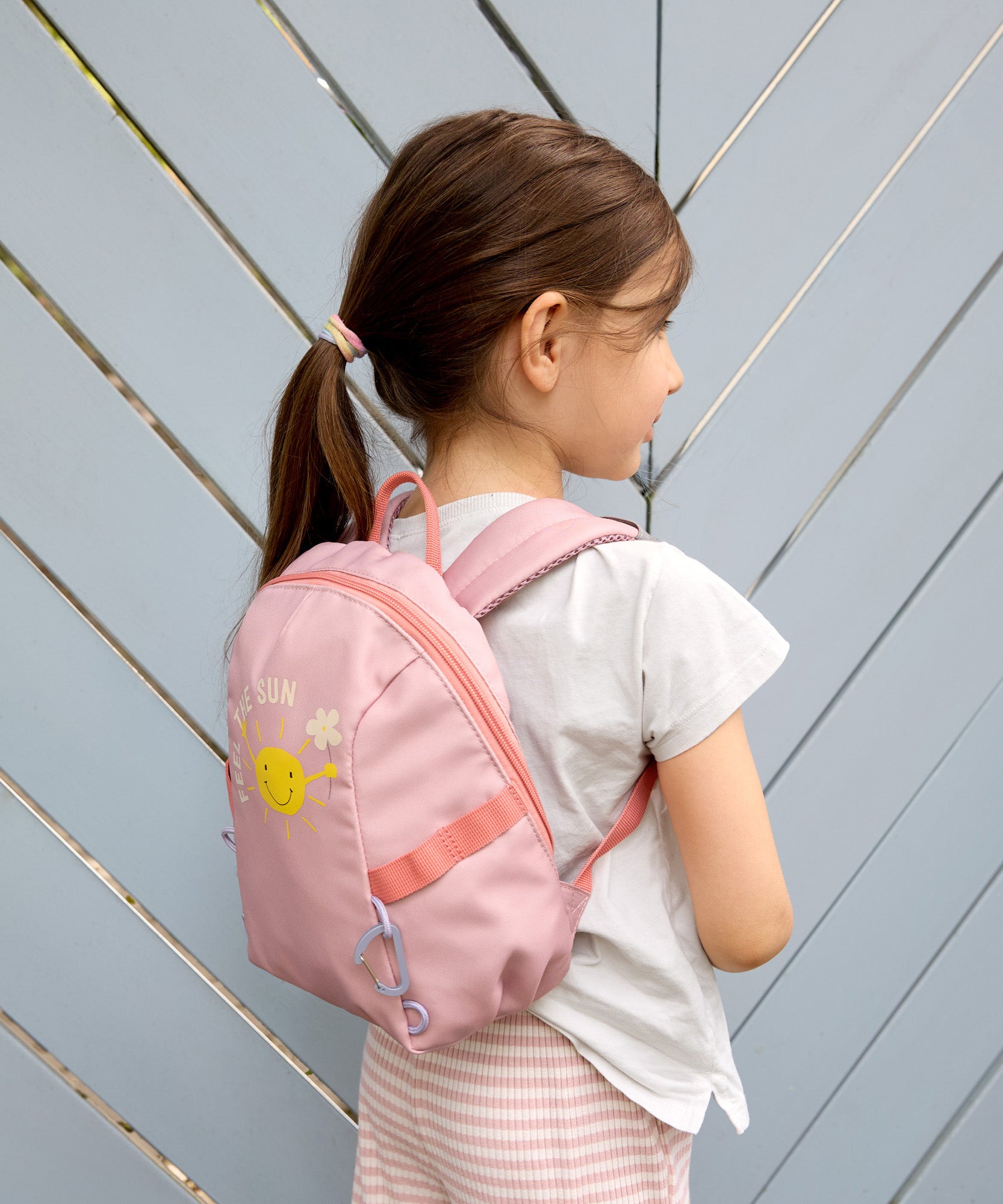 A girl wearing the Lassig tiny backpack for toddlers and little kids, in a light pink with a sunshine face cartoon.