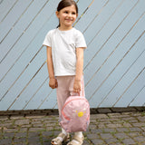 A little girl holding the Lassig tiny backpack for toddlers and kids, in a light pink.