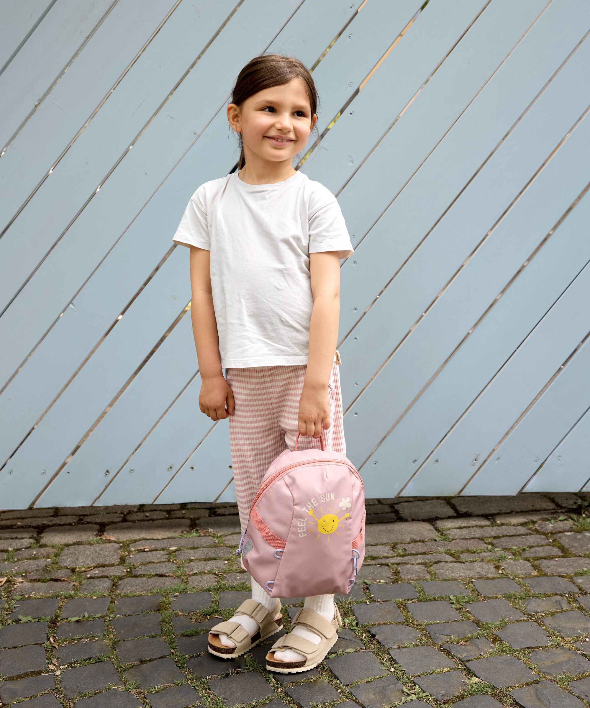 A little girl holding the Lassig tiny backpack for toddlers and kids, in a light pink.