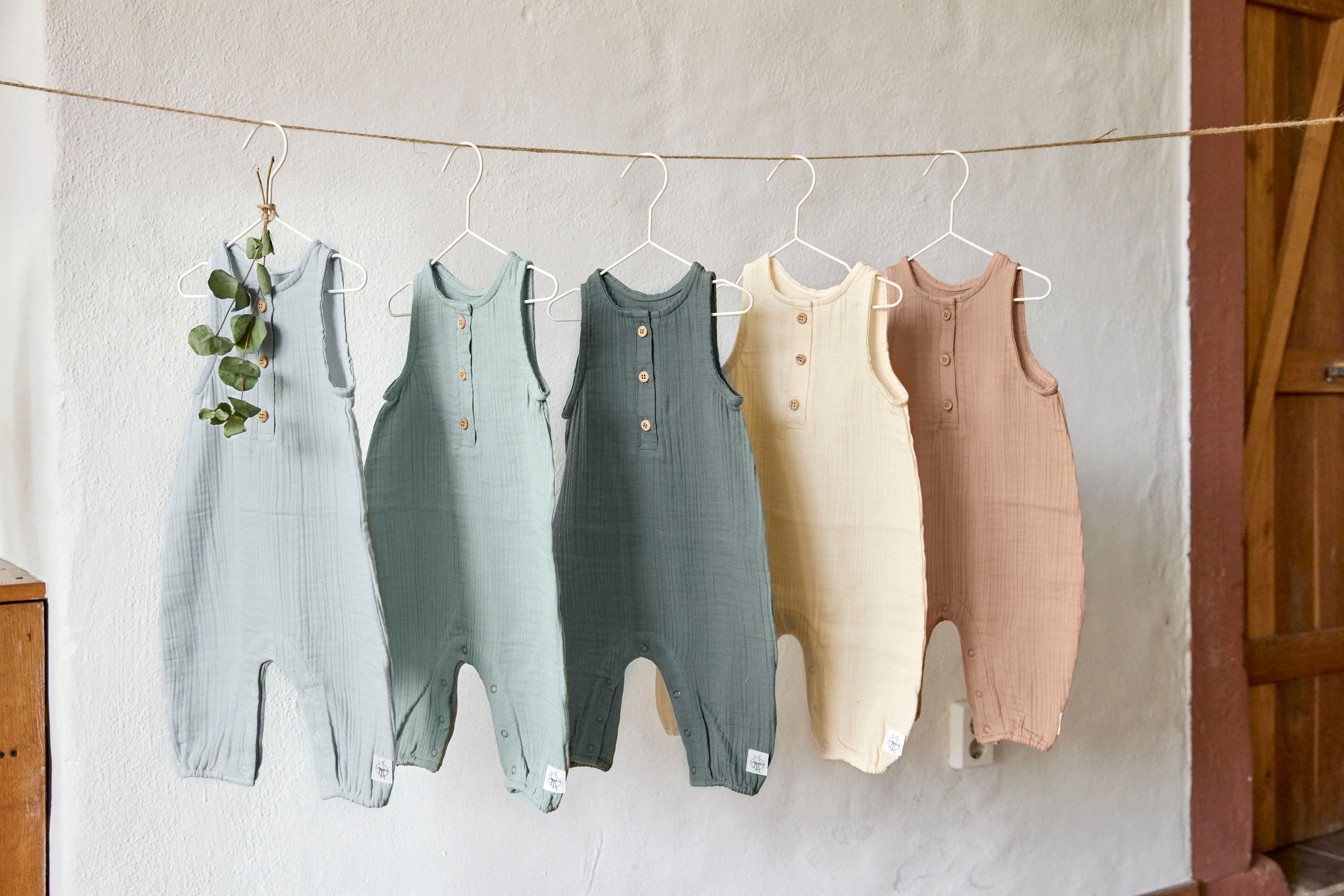 5 different coloured Lassig organic cotton muslin jumpsuits, hanging on a string of twine.