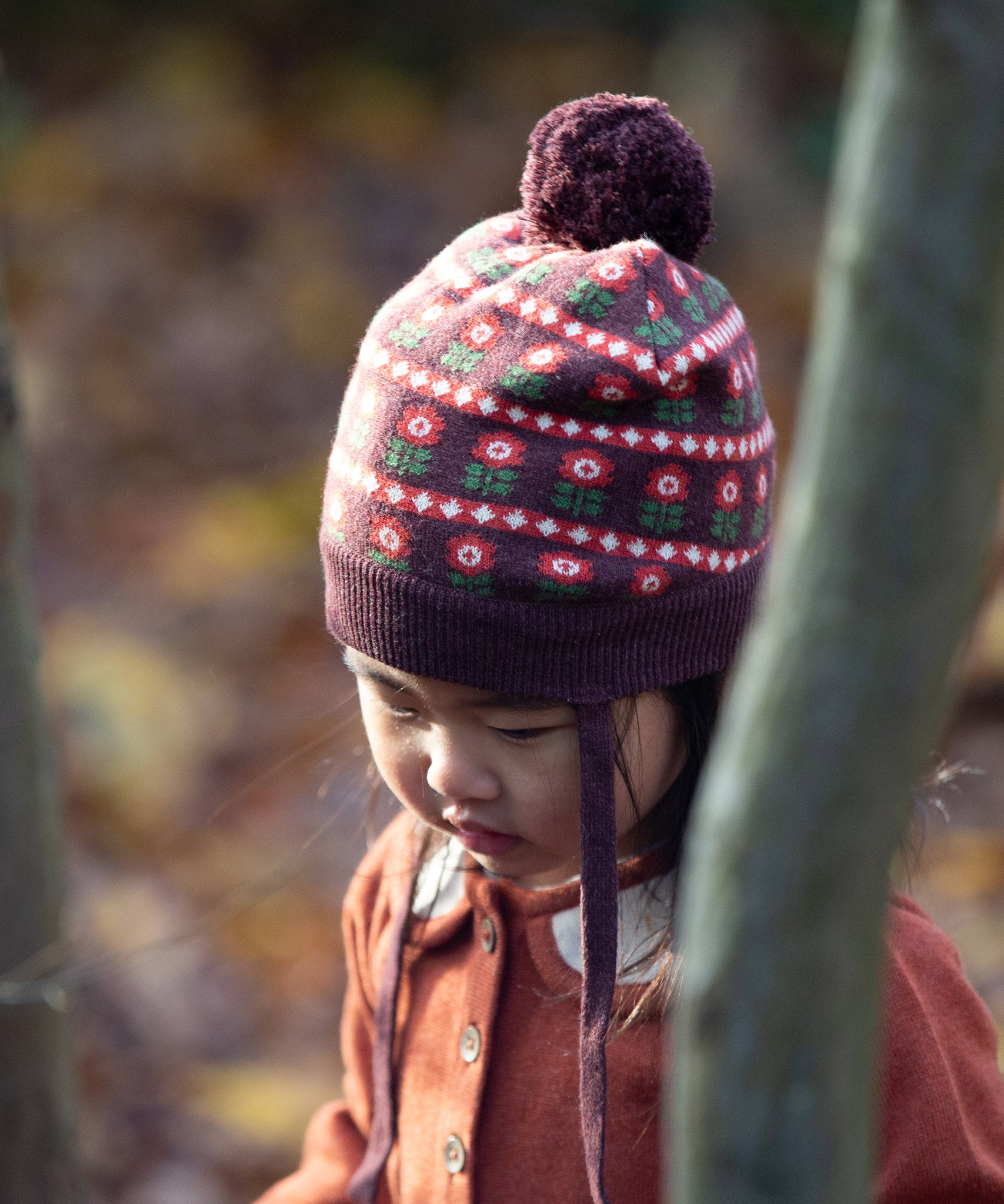 Close up of baby wearing a cosy chocolate brown knit hat with a floral pattern, brown pom pom and chin straps, made from Fairtrade GOTS organic cotton by Little Green Radicals, shop now at Babipur.