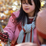 Blue and cream stripes adorn the knitted scarf worn by a child in the forest — designed by Little Green Radicals, produced from Fairtrade and GOTS-certified organic cotton, sold at Babipur.