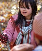 Blue and cream stripes adorn the knitted scarf worn by a child in the forest — designed by Little Green Radicals, produced from Fairtrade and GOTS-certified organic cotton, sold at Babipur.