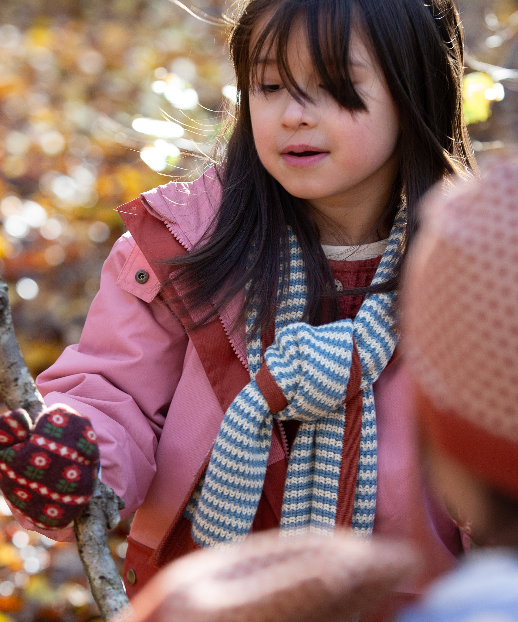Blue and cream stripes adorn the knitted scarf worn by a child in the forest — designed by Little Green Radicals, produced from Fairtrade and GOTS-certified organic cotton, sold at Babipur.