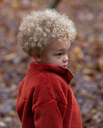 Child playing in a forest wearing Polar Fleece Jacket in Ginger red with yellow zipped pockets by Little Green Radicals, made from recycled fleece, buy now at Babipur.