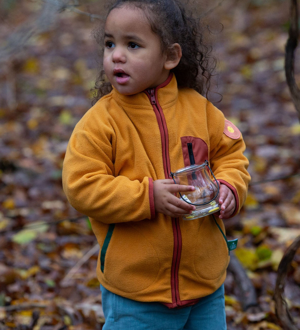 Child playing in a forest wearing a yellow gold zipped fleece jacket from Little Green Radicals, made from recycled polar fleece, for sale at Babipur.