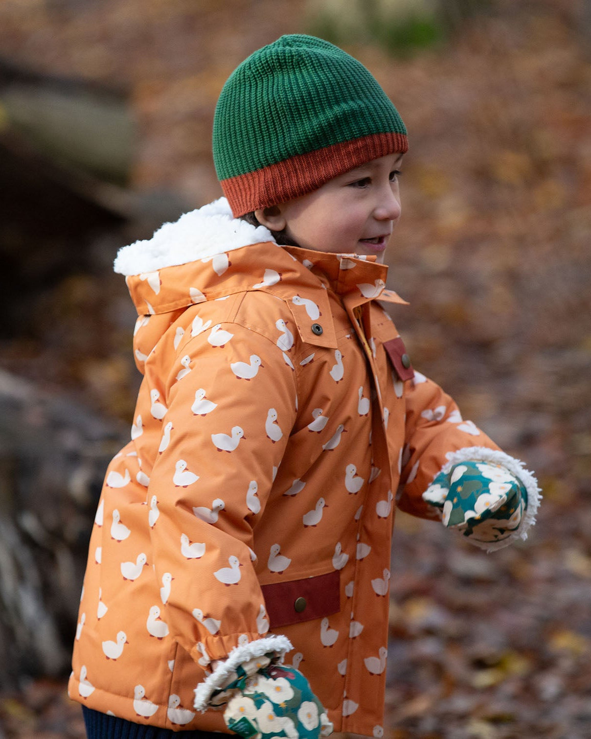 Side view of a child running in a forest wearing a hooded Little Green Radicals Waterproof Winter School Coat, with elasticated sleeves, made from recycled bottles, with a yellow duck print, for sale at Babipur.