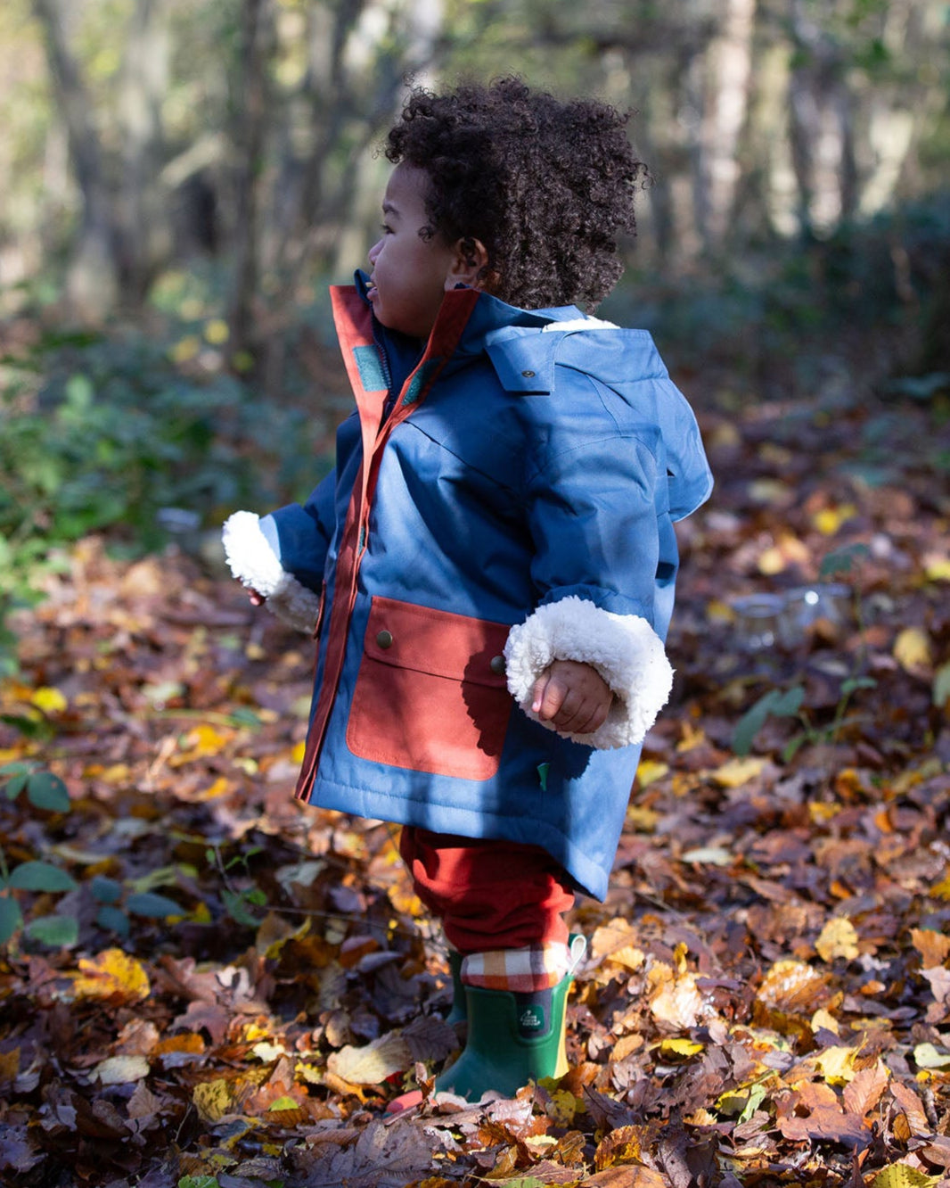 Baby in a forest wearing a hooded Little Green Radicals Waterproof Winter School Coat, with elasticated sleeves, made from recycled bottles, with a Moonlight Blue print with large pockets in deep red, for sale at Babipur.