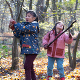 Girl stood in a forest wearing and Waterproof (5000M rating) and warm, PVC free, a cosy hooded kids winter coat by Little Green Radicals, in a Night Moon blue print, with red pockets, available now at Babipur.