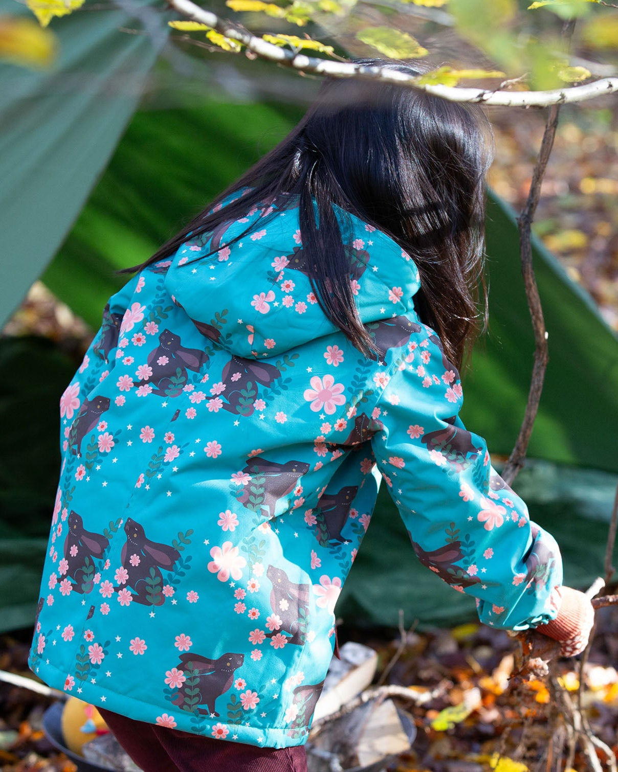Back of girl playing in a forest wearing a hooded Little Green Radicals Waterproof Winter Coat, made from recycled bottles, with a turquoise Winter Rabbits print, for sale at Babipur.