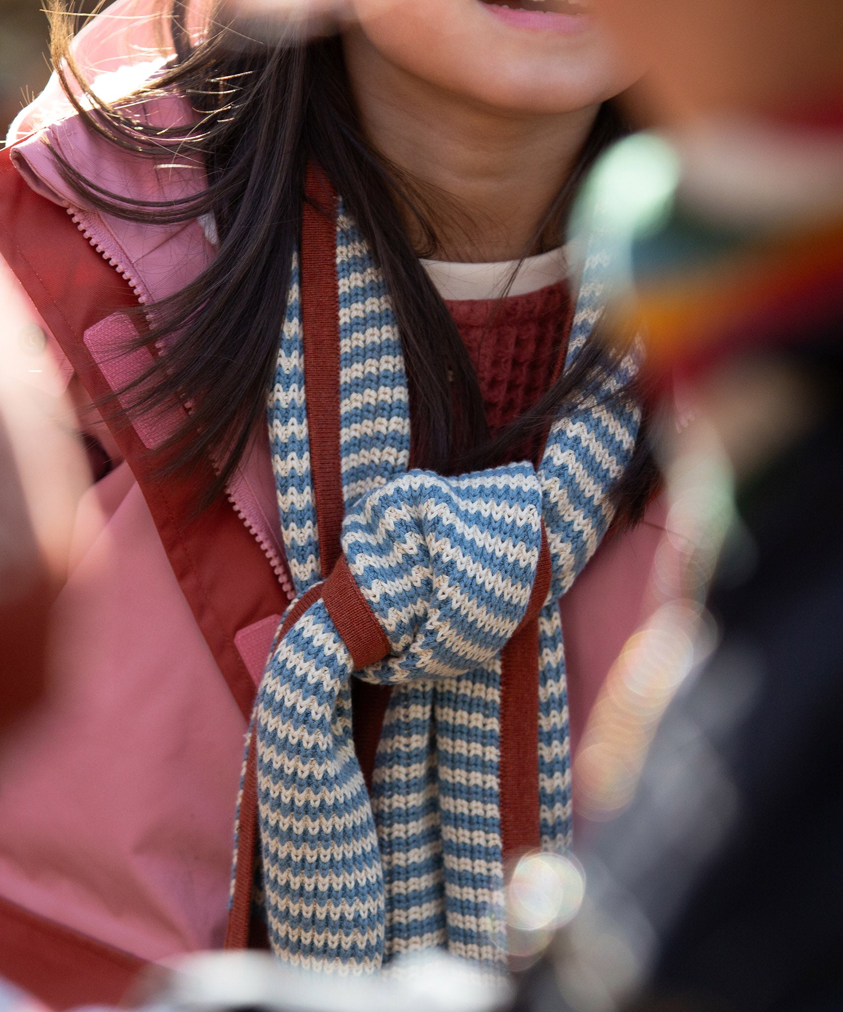 Close up of a child in the forest wrapped in a blue and cream striped knitted scarf from Little Green Radicals, crafted with Fairtrade and GOTS-certified organic cotton, available at Babipur.