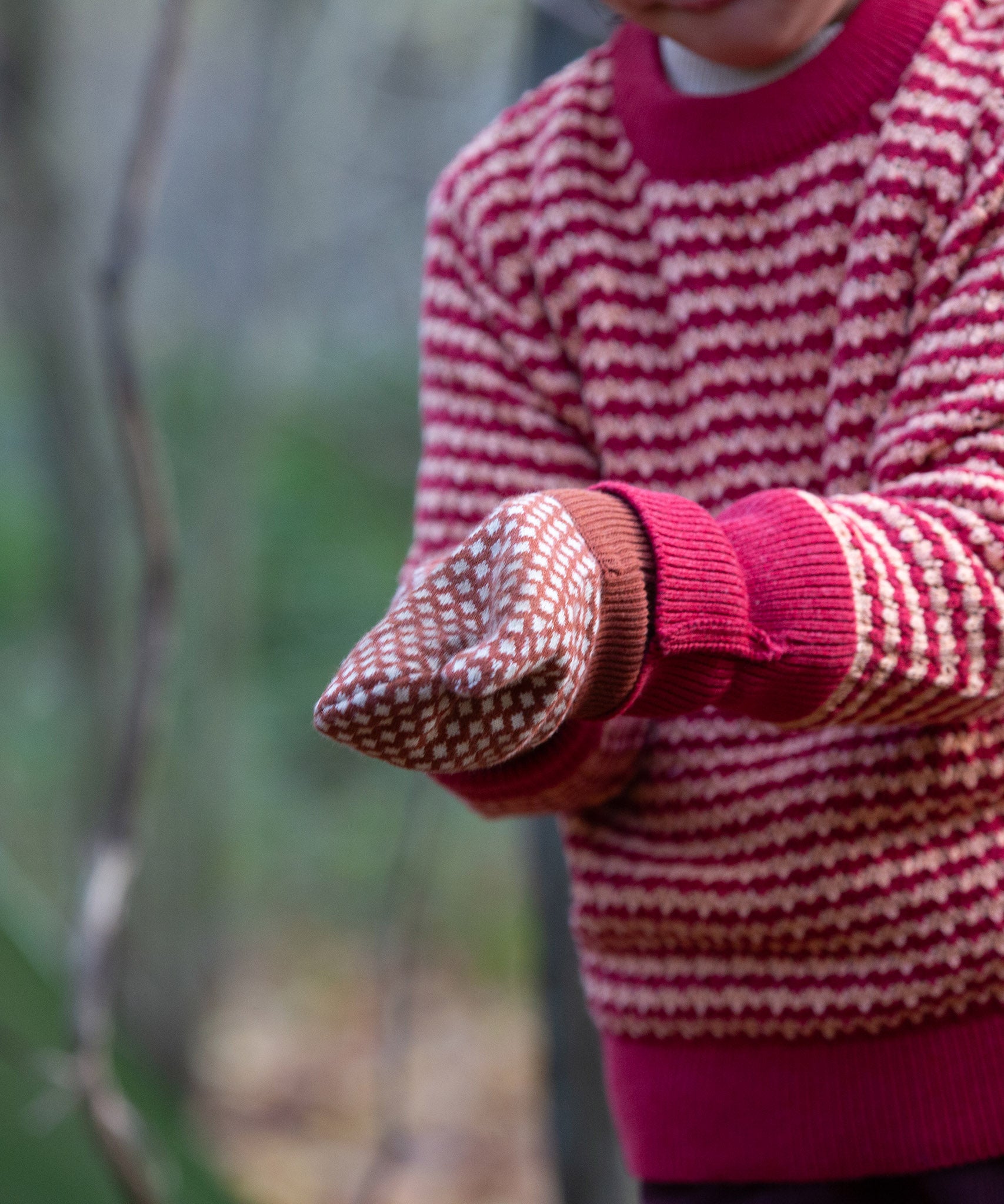 Close up of a child wearing a pair of Little Green Radicals Knitted Mittens with a Ginger Orange and Cream Diamond Pattern and thick orange cuffs - made from soft Fairtrade certified and GOTS organic cotton, for sale at Babipur.