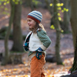 Child stood in a forest wearing a blue and cream striped Knitted Beanie by Little Green Radicals with a red trim - made from soft GOTS organic and Fairtrade certified cotton - for sale at Babipur.