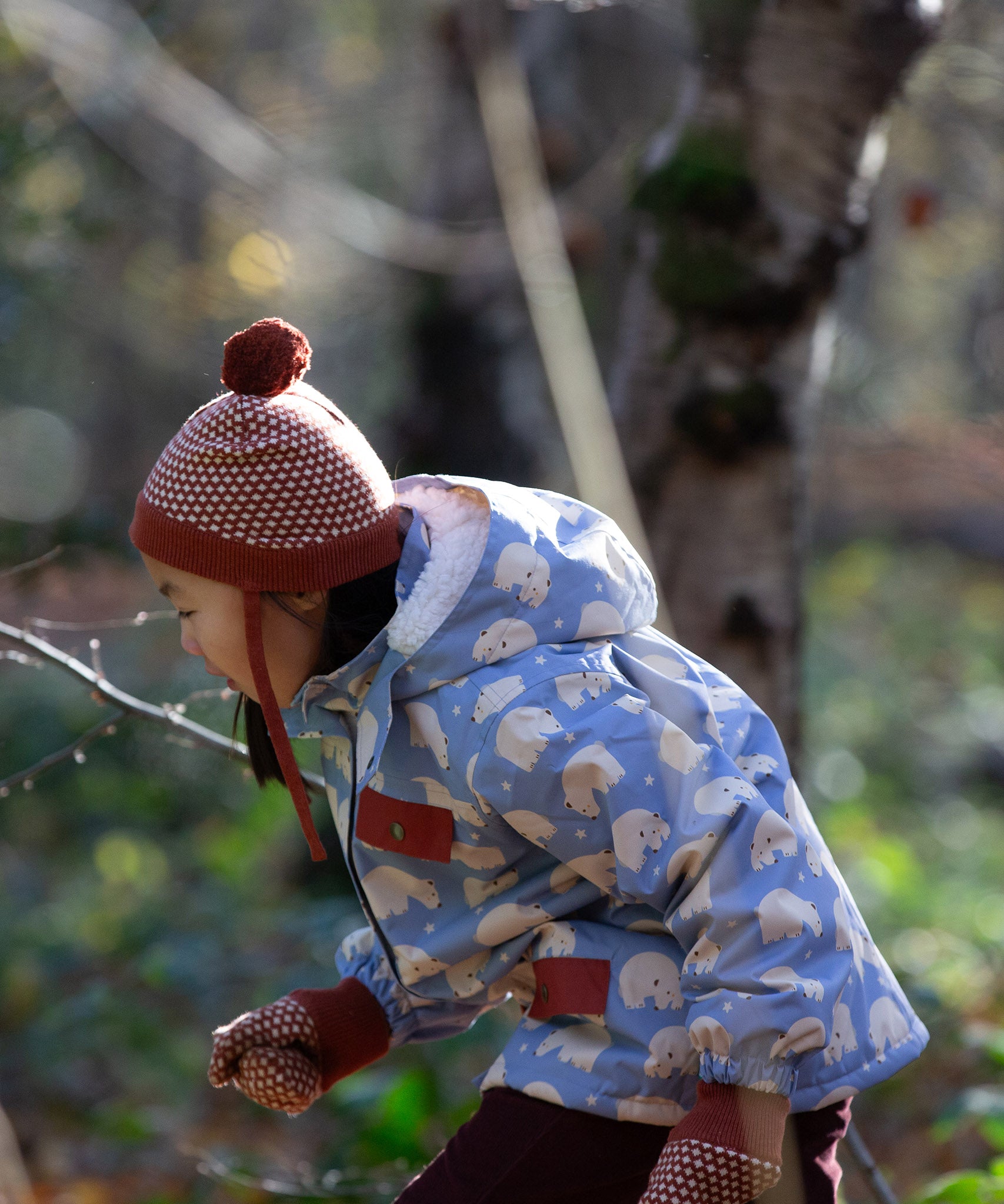 Side of child in a forest wearing a Little Green Radicals knitted ginger orange hat with a diamond pattern and a deep crimson pom pom, made from soft Fairtrade and GOTS certified organic cotton - available now at Babipur.