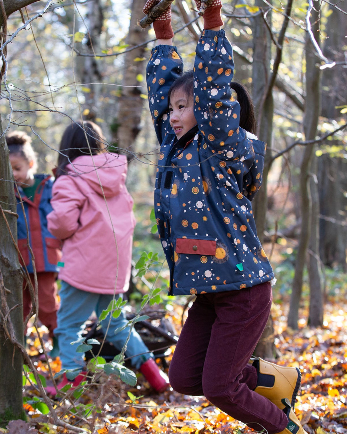 Girl hanging on a tree in a forest, wearing a hooded Little Green Radicals Waterproof Winter School Coat, made from recycled bottles, with a navy night moon print, for sale at Babipur.