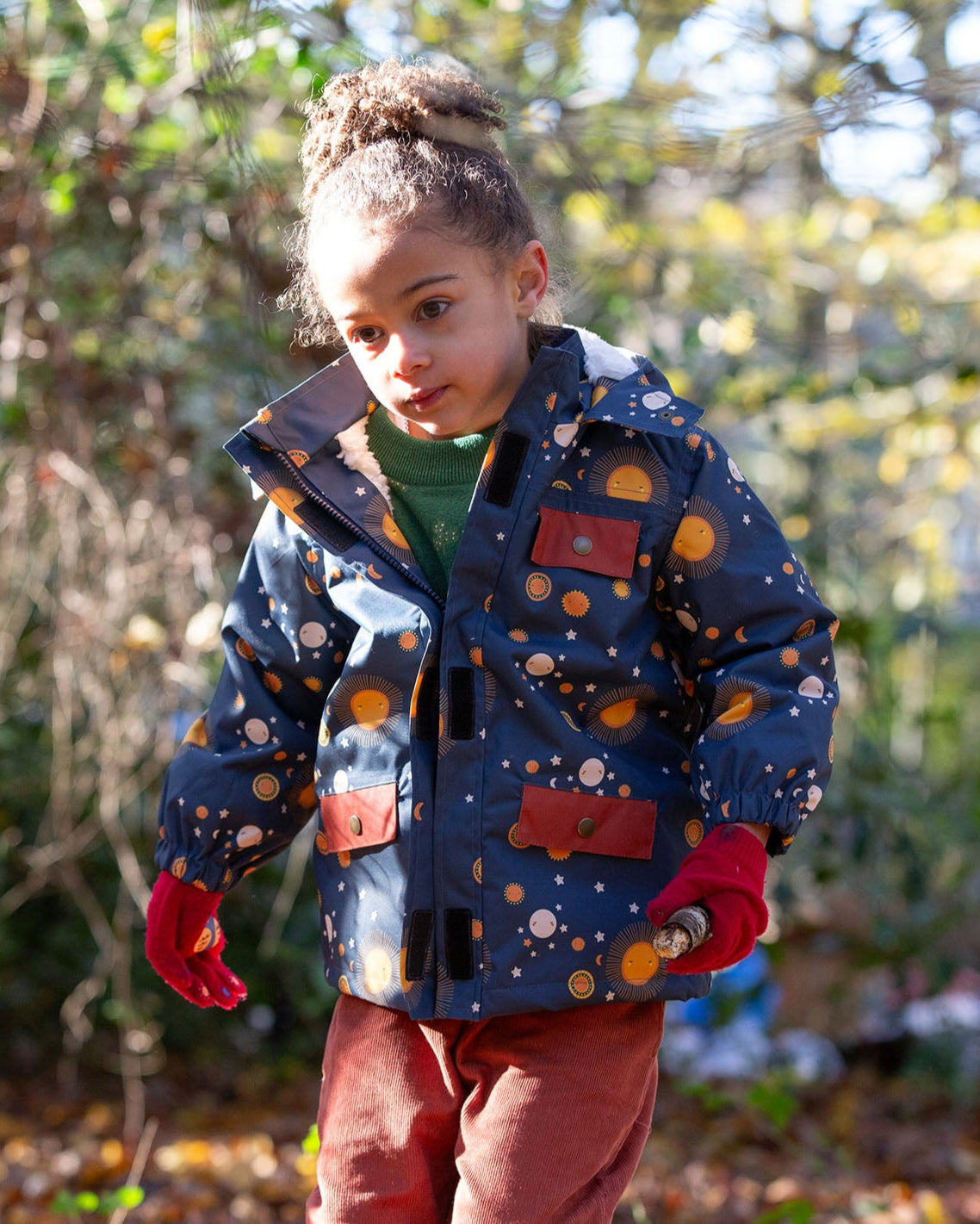 Girl stood in a forest wearing a Warm and Waterproof (5000M rating), PVC free kids school coat by Little Green Radicals, in a navy night moon print, with red front pockets, buy now at Babipur.