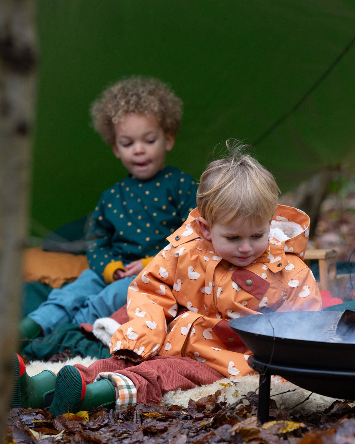 Boy sat at a campfire wearing a Little Green Radicals Waterproof Winter Coat with a cosy fleece hood, made from recycled bottles and a cosy polyester fleece, in a gold duck print, available now at Babipur.