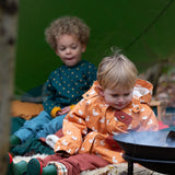 Boy sat at a campfire wearing a Little Green Radicals Waterproof Winter Coat with a cosy fleece hood, made from recycled bottles and a cosy polyester fleece, in a gold duck print, available now at Babipur.