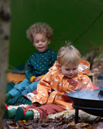 Boy sat at a campfire wearing a Little Green Radicals Waterproof Winter Coat with a cosy fleece hood, made from recycled bottles and a cosy polyester fleece, in a gold duck print, available now at Babipur.