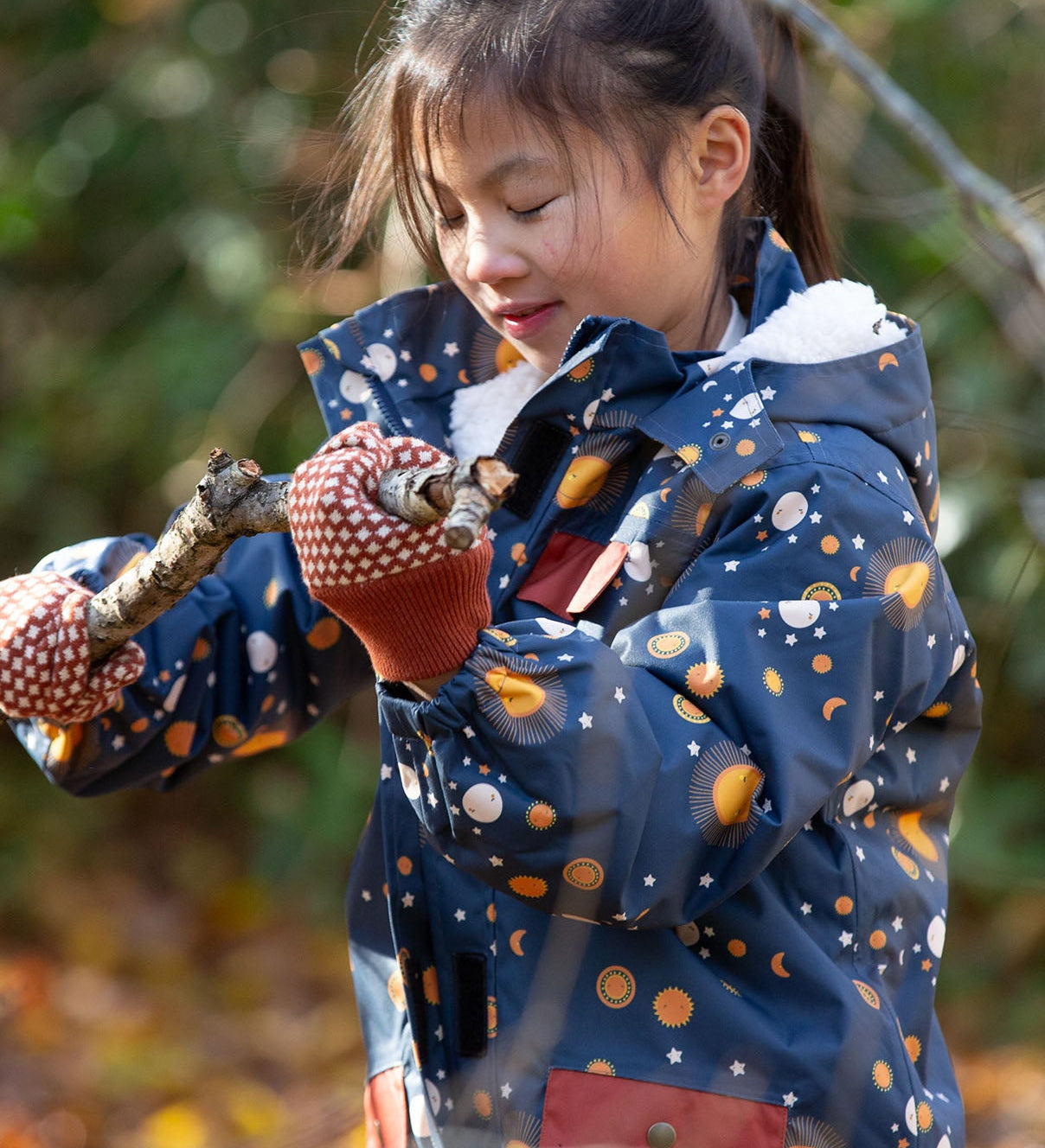 Child in a forest wearing a Little Green Radicals Waterproof Winter Coat in a navy Night Moon print, with a cosy fluffy hood, made from recycled bottles and a cosy polyester fleece, for sale at Babipur.