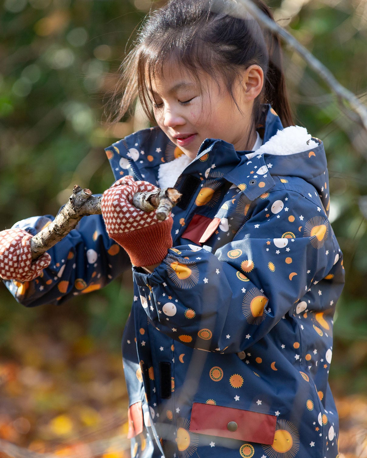 Child in a forest wearing a Little Green Radicals Waterproof Winter Coat in a navy Night Moon print, with a cosy fluffy hood, made from recycled bottles and a cosy polyester fleece, for sale at Babipur.