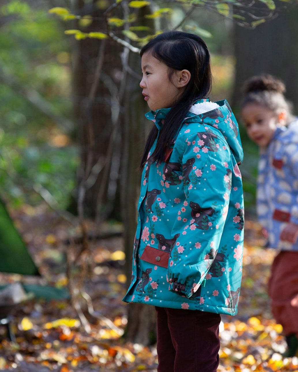 Child in a forest wearing a Little Green Radicals Waterproof Winter Coat in a turquoise Winter Rabbits print, with a cosy fluffy hood, made from recycled bottles and a cosy polyester fleece, for sale at Babipur.