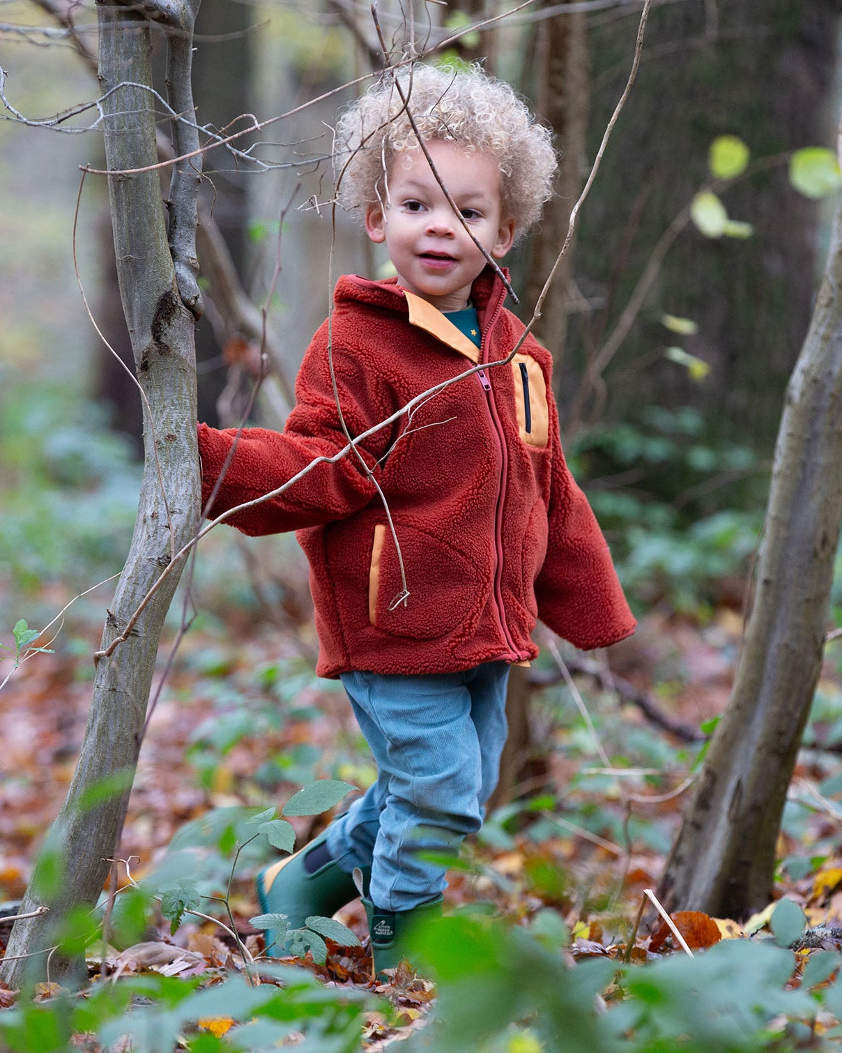 Child stood in a forest wearing an organic red ginger zipped fleece jacket with a big yellow pocket from Little Green Radicals, made from recycled polar fleece, available now at Babipur.