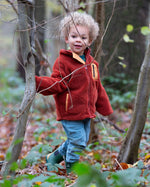 Child stood in a forest wearing an organic red ginger zipped fleece jacket with a big yellow pocket from Little Green Radicals, made from recycled polar fleece, available now at Babipur.