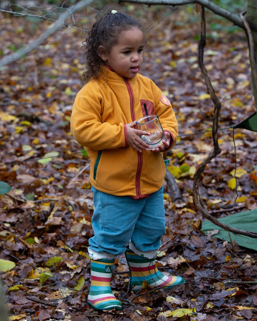 Child stood in a forest wearing an organic yellow gold zipped fleece jacket with a big red pocket from Little Green Radicals, made from recycled polar fleece, available now at Babipur.