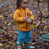 Child stood in a forest wearing an organic yellow gold zipped fleece jacket with a big red pocket from Little Green Radicals, made from recycled polar fleece, available now at Babipur.