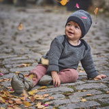 A child sitting on a cobbled surface with falling Autumn leaves and wearing the Mamalila quilted Berlin booties in Khaki available at Babipur.