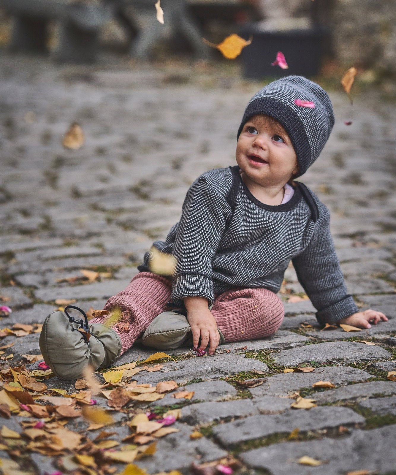 A child sitting on a cobbled surface with falling Autumn leaves and wearing the Mamalila quilted Berlin booties in Khaki available at Babipur.