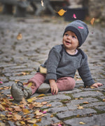 A child sitting on a cobbled surface with falling Autumn leaves and wearing the Mamalila quilted Berlin booties in Khaki available at Babipur.