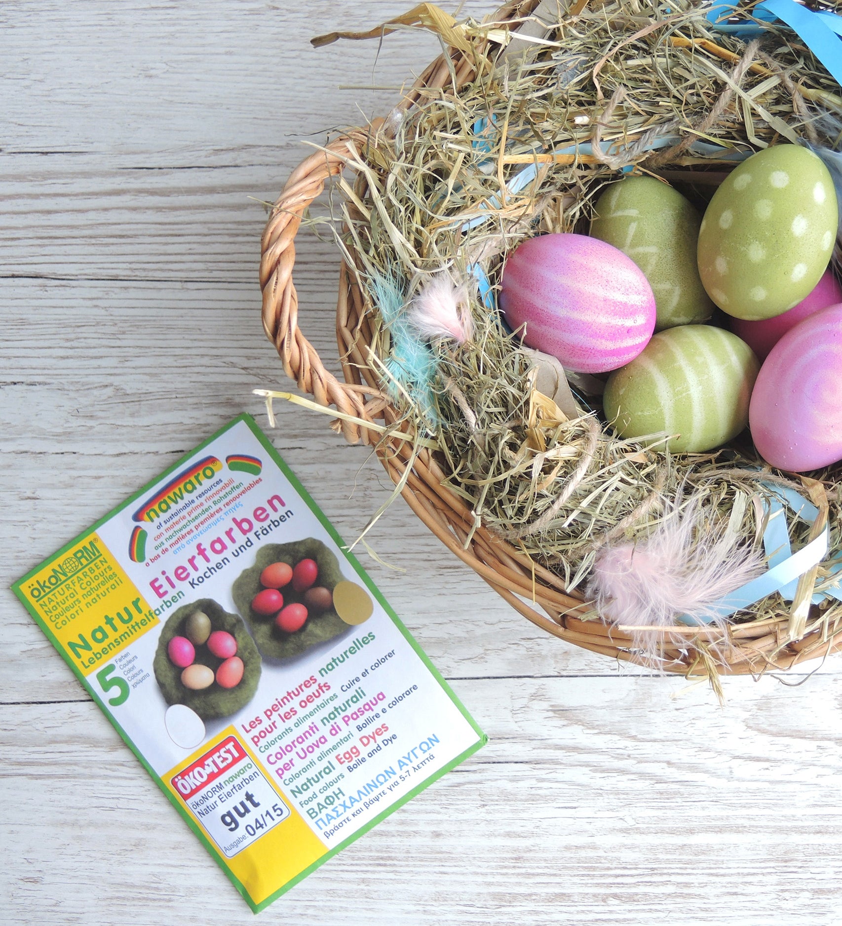 ÖkoNORM nawaro natural egg dye set placed on a light wooden surface beside a wicker basket filled with hay and dyed Easter eggs. The packaging is green and white, featuring an image of naturally dyed eggs in shades of red, pink, yellow, and brown. The label highlights that the dyes are made from natural ingredients. The basket contains decorated eggs in pastel shades of pink and green, some with polka dot and striped patterns, nestled among hay, feathers, and blue ribbon
