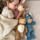 A girl sleeping and holding onto a Olli Ella cozy dinkum bunny pickle doll with 2 other bunny dinkums on the bed. These fluffy toy dolls are available at Babipur
