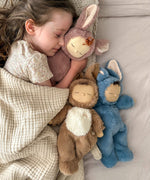 A girl sleeping and holding onto a Olli Ella cozy dinkum bunny pickle doll with 2 other bunny dinkums on the bed. These fluffy toy dolls are available at Babipur