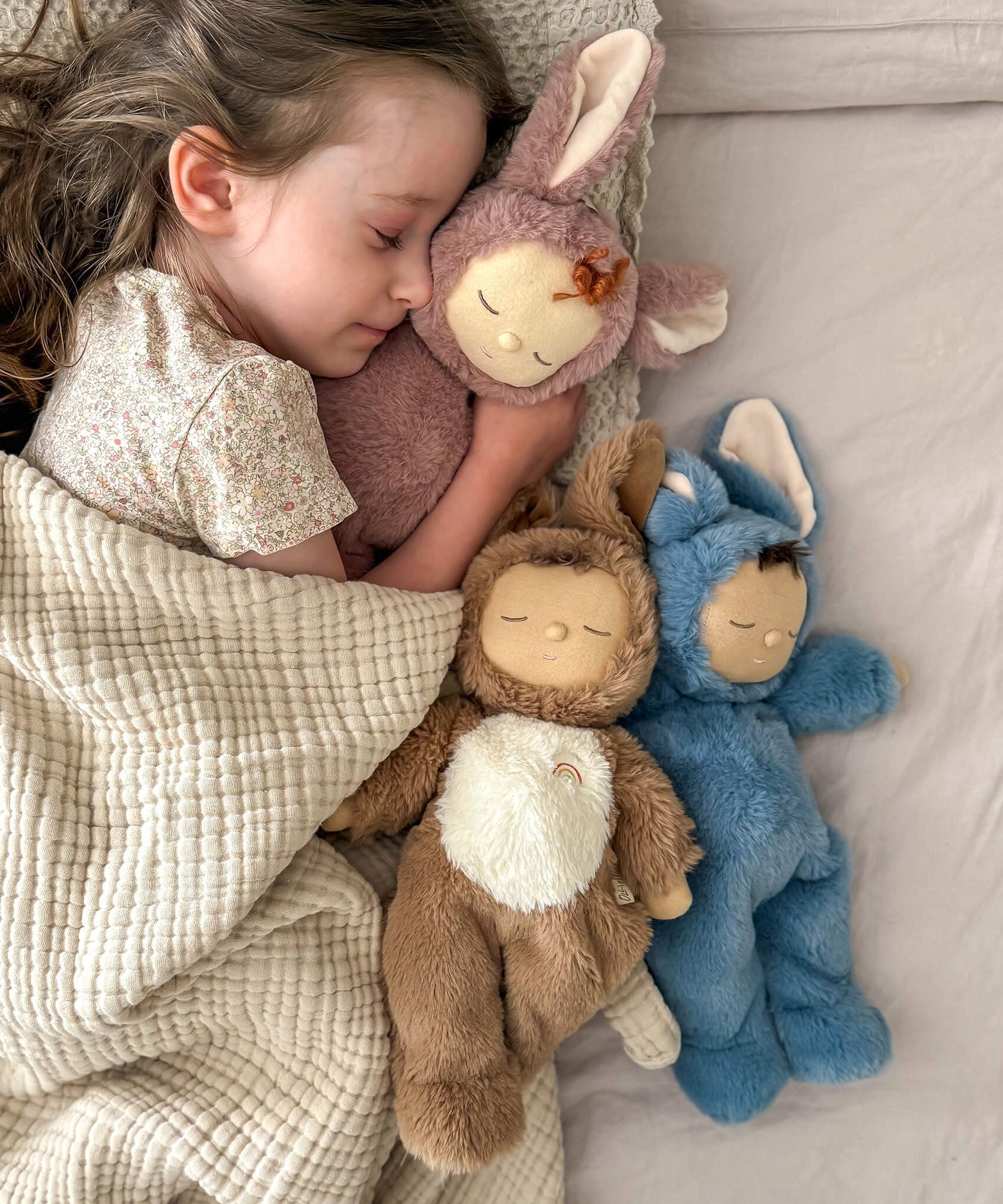 A girl sleeping and holding onto a Olli Ella cozy dinkum bunny pickle doll with 2 other bunny dinkums on the bed. These fluffy toy dolls are available at Babipur