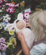 A little girl holding a Olli Ella cozy dinkum bunny pip doll surrounded by flowers. This is a soft and fluffy doll with a sleeping face, cute floppy ears and available at Babipur.