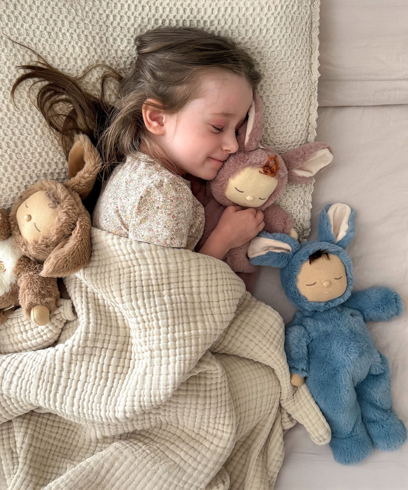 A girl sleeping next to Olli Ella cozy dinkum bunnies including the blue twiggy bunny. These fluffy toy dolls are available at Babipur
