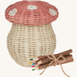 An Olli Ella Porcini mushroom shaped Basket with a pink spotted lid, and wooden twig colouring pencils wrapped together with string next to it pictured on a cream background